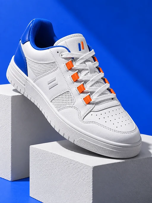 Ein weißer Sneaker steht auf weißen Architekturblöcken vor kräftigem Blau. Feine Lederdetails und klare Schatten bestimmen das Studiofoto.