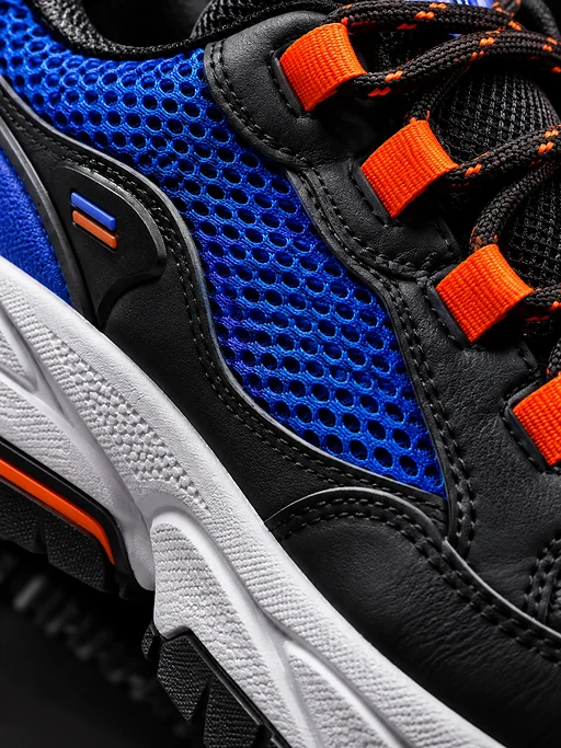 Die Makroaufnahme zeigt blaues Mesh, orangefarbene Schnürösen, schwarzes Leder und die strukturierte Sohle eines Sneaker.